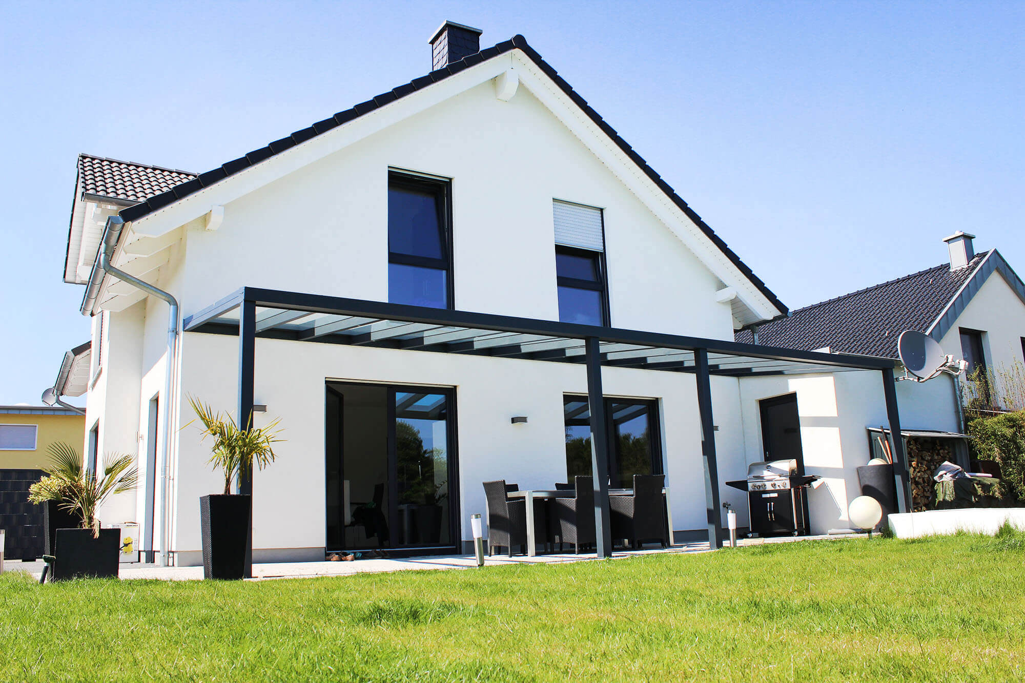 Terrassenüberdachung schwarz mit durchsichtigem VSG-Glas an weißem Haus mit 2 Stützen in der Mitte